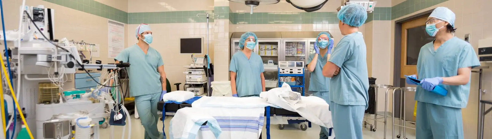 Group of medical professionals in light blue scrubs and surgical caps standing around a hospital bed.