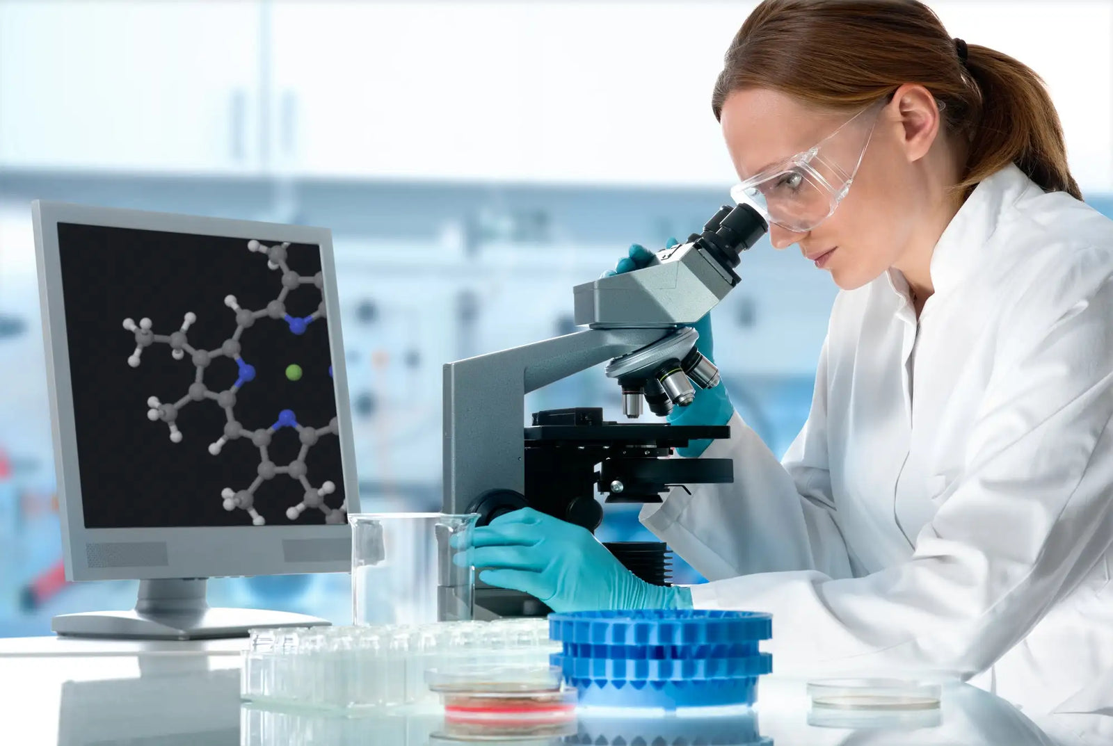 A sleek, metallic microscope with black accents and adjustable lenses sits on a lab bench beside a computer monitor displaying molecular structures.