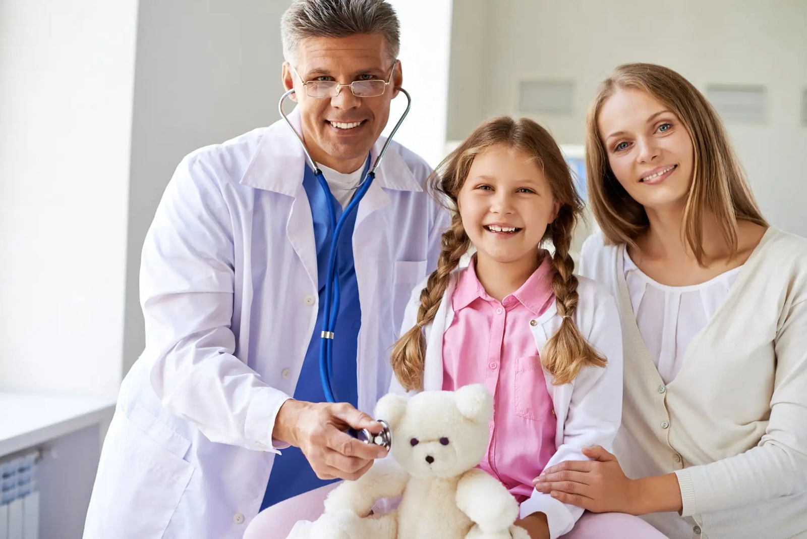 A white plush teddy bear with black button eyes and a stitched nose, held by a smiling girl in a pink shirt.