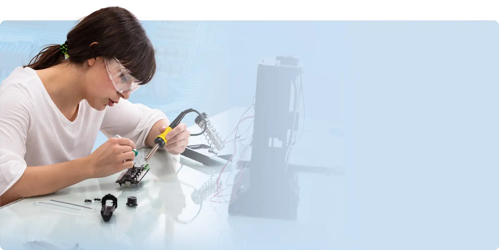 Woman wearing safety goggles and a white shirt, soldering an electronic circuit board with a yellow-handled soldering iron