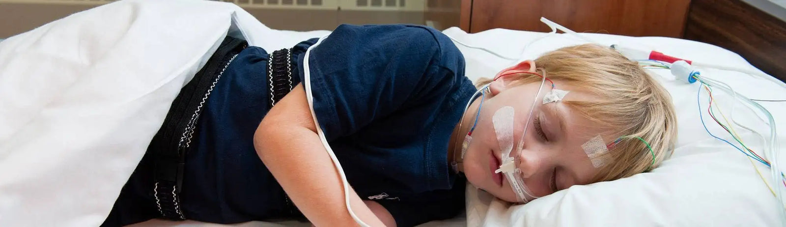 A young child with blonde hair lies in a hospital bed, wearing a navy blue shirt and connected to medical monitoring equipment.