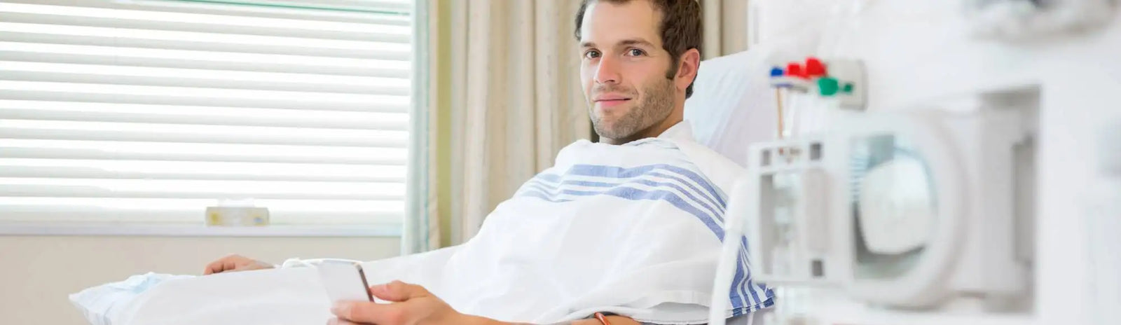 A man in a hospital gown with blue and white stripes sits on a bed, holding a pen.