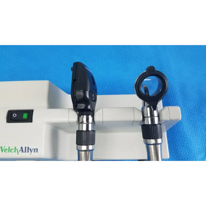 Black and silver welch allyn otoscope with circular lens and angled head mounted on white wall transformer base unit for reuse medical use