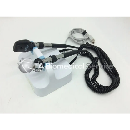 Black and silver welch allyn green series otoscope with coiled cable resting on white stand for wall transformer use